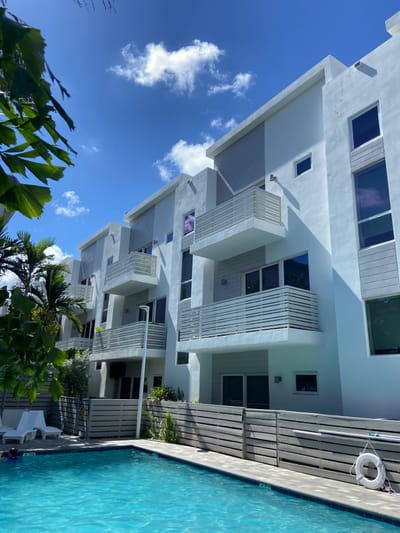 The image displays an outdoor scene in Pompano Beach, United States, featuring a modern residential complex and a swimming pool under a bright blue sky with scattered white clouds. The foreground contains a clear blue swimming pool bordered by light grey tiles. To the left of the pool, two white poolside lounge chairs are visible. A horizontally slatted, light grey fence separates the pool area from the multi-story residential buildings. A white life preserver with red markings is suspended on this fence at the right side of the pool.
The residential structures are characterized by a contemporary design, primarily with white or light grey exterior walls and sections featuring horizontal grey siding. Multiple windows, some displaying purple reflections, are present across the facades. Each visible unit includes at least one balcony equipped with white horizontal slatted railings. A tall, slender grey outdoor light fixture stands upright between the fence and the buildings. Green tropical foliage, including palm trees, is visible behind the fence and in the upper left foreground, partially obscuring the view. One individual is partially visible in the lower-left section of the swimming pool, with only their head and shoulders visible above the water surface. The scene is illuminated by strong sunlight, casting shadows from the buildings.