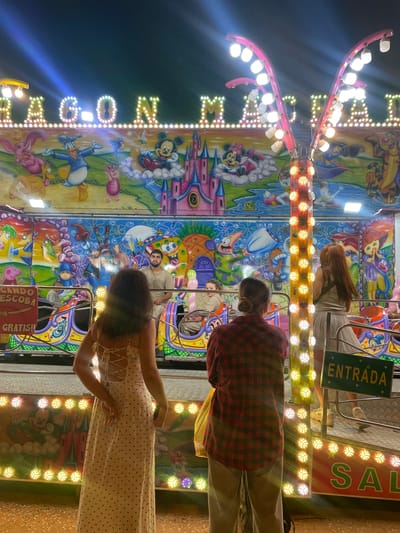 This is an outdoor nighttime photograph taken at a fairground in Canet de Mar, Spain. The image features a colorful amusement ride decorated with cartoon characters, including Mickey Mouse, Donald Duck, and characters from "The Little Mermaid" and "SpongeBob SquarePants." The ride is illuminated by strings of bright lights. In the foreground, three individuals are visible from behind, appearing to be waiting for or exiting the ride. One person on the left wears a white polka-dot dress, another in the center wears a red and black plaid shirt and light-colored pants, and a third person on the right wears a light-colored dress. A green sign with the word "ENTRADA" (Entrance) is visible to the right. The ground appears to be sandy with some scattered confetti.