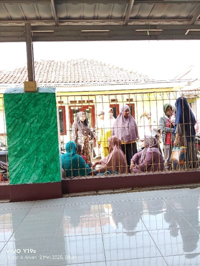 The image shows a group of nine women standing and sitting outside a home in Kecamatan Anjatan, Indonesia, at the coordinates -6.419, 107.98. The women are wearing traditional Islamic clothing. They are standing in a line behind a fence, and most are facing the camera. There is a green wall on the left side of the image, and the ground is covered in tiles. The home has a tiled roof. A metal fence surrounds the home and the women stand behind it. The women are all interacting with each other and appear to be engaged in conversation. The image was taken in daylight with bright conditions. The image was taken on a sunny day, and the sky is bright. The sun is shining on the scene and there is no sign of clouds. The image appears to be of a gathering of women in a home in Kecamatan Anjatan, Indonesia. There is a large green wall to the left of the image, and a tiled area to the right of the image. The women are all of varying ages and body types, and appear to be dressed for a special occasion.