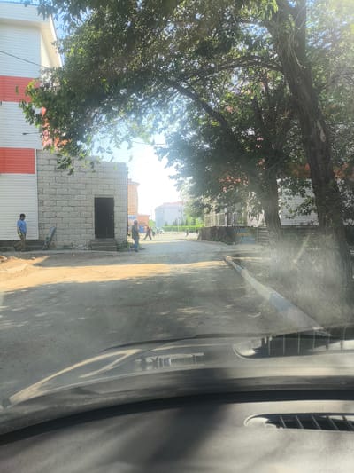 The image, taken from inside a vehicle, depicts a street scene in the city of Abay, Kazakhstan, during daylight. In the foreground, the dashboard and windshield of the vehicle are visible. The street itself is unpaved and dusty, with shadows from trees cast across it.
On the left side of the street, a two-story building is present. The lower part of this building is constructed with light-colored brickwork, featuring a dark doorway and a set of wooden steps leading up to it. Above the brickwork, the building's facade is clad in horizontal white and orange panels. A person in a blue shirt and beige pants stands to the left of this building, facing away from the camera and towards the street.
Further down the street, on the right side, a large tree with a thick trunk and leafy branches arches over the road, partially obscuring the sky and sunlight. Beyond the tree, other buildings are visible, including a structure with large windows and a colorful base, and in the distance, several other buildings with white exteriors and darker roofs.
Several individuals are present further down the street. Two people appear to be walking or working near a building on the right. In the middle distance, two more figures are walking along the street, and further still, a group of people are visible, with a car driving away from the camera in the background. The overall atmosphere suggests a residential or commercial area undergoing some activity, possibly construction or maintenance, given the presence of the unpaved road and the people engaged in various actions. The lighting indicates it is a sunny day, with the sun creating lens flare in some parts of the image.