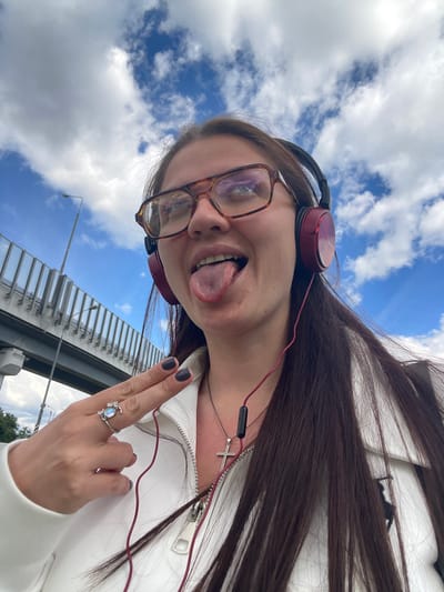 A young woman is pictured in an upward-facing selfie. She is wearing glasses, headphones, a white jacket, and a silver necklace with a cross pendant. Her hair is long and brown. She is sticking her tongue out and displaying a ring on her finger. The background consists of a blue sky with white clouds, a bridge, and a few light poles. Given the location information, the image was taken near Razvilka, Russia.