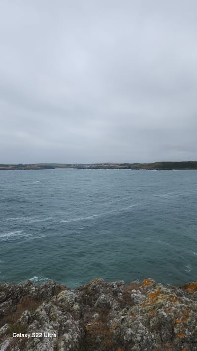 The image depicts a choppy, dark blue sea under a heavily overcast sky. In the distance, a low-lying coastline is visible, dotted with houses and some wind turbines. The foreground is dominated by a rugged, rocky cliff face covered in lichen and dry vegetation. The scene appears to be located on the coast of Cemaes, United Kingdom, given the presence of the "Galaxy S22 Ultra" watermark which is associated with mobile phone imagery. No people or specific actions are visible in the image.