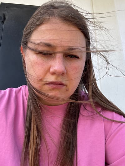In Uyutnoye, Ukraine, a young woman with long brown hair, partially obscuring her face due to the wind, poses for a selfie. She is wearing a pink crew-neck t-shirt. The background consists of a dark, possibly metal, surface on the left and a light-colored, textured wall on the right. Her expression is neutral, with her eyes looking down and slightly closed. The image captures the woman from the chest up, emphasizing the impact of the wind on her hair.
