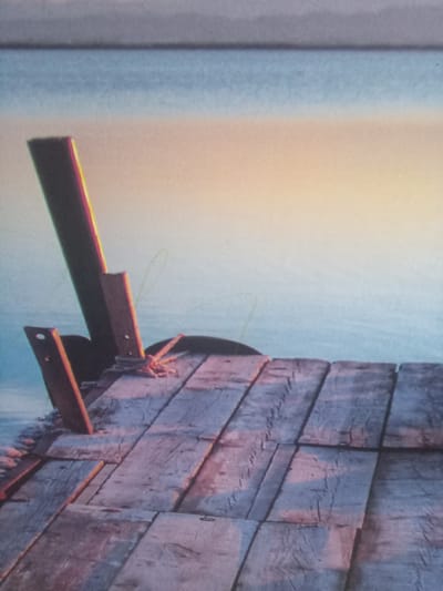The image shows a close-up view of wooden planks forming a pier or dock extending over a body of water. The wood appears weathered and worn, with visible grain and some discoloration from age and exposure. There are two dark, upright wooden posts on the left side of the frame, one taller than the other, with metal brackets attached to them. A rope is loosely tied around one of the posts. The water is calm and reflects the soft colors of the sky, which transitions from a pale blue at the horizon to warmer tones of yellow and pink near the bottom of the frame, suggesting either dawn or dusk. The overall scene evokes a sense of quiet stillness and natural beauty. There are no people or animals visible in the image. The photograph is taken from a low angle, looking out over the water from the perspective of the dock. The location in Terebovlia, Ukraine, is not discernible from the visual information provided in the image.