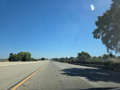 This media file captures a view from a vehicle driving on a highway in Sacramento, California. The scene is set on a sunny day with a clear blue sky, indicating good weather. The highway stretches into the distance, with multiple lanes of concrete pavement marked by yellow and white lines. Several vehicles, including cars and trucks, are visible on the road, suggesting moderate traffic.
To the left of the highway, a concrete barrier separates the lanes from an adjacent area with a few scattered vehicles and a line of green trees. To the right, a metal guardrail runs alongside the road, separating it from a residential area with houses and more lush greenery. A large tree with dark green foliage is prominently featured on the right side of the frame, with its branches extending towards the sky.
The time of day appears to be late morning or early afternoon, given the bright sunlight and the shadows cast on the road. There are no people clearly visible in the media file. No text is discernable in the image. The overall impression is one of a typical day of travel on a suburban or exurban highway.