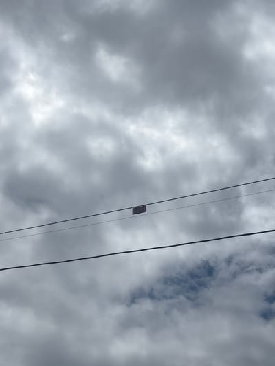 The image captures a cloudy sky above Bo Y, Vietnam. Two power lines stretch horizontally across the frame. Attached to the upper power line is a small, dark-colored rectangular object, possibly a tag or a piece of debris. The clouds are dense and gray, obscuring most of the sky, with hints of blue peeking through in some areas. No people or vehicles are visible in the scene.