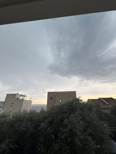 The image depicts an exterior scene from what appears to be a balcony or elevated vantage point, looking upwards and outwards. Dominating the left side of the frame is a large, dark, angled structure, possibly a wall or railing, partially obscuring the view. The sky is filled with dramatic, heavy clouds, suggesting an overcast or stormy weather condition.
In the mid-ground, there are portions of buildings. One building, with a red-tiled roof and beige exterior, is partially visible on the right side, towards the top. Below and to the right of that, a more modern, beige building with multiple windows and a balcony is seen. In the distance, beyond the buildings and a dense olive tree, a body of water, likely the sea, is visible under the hazy sky, with the horizon faintly discernible.
The time of day appears to be either early morning or late afternoon, given the diffused light and the color of the sky, which transitions from a pale yellow near the horizon to a muted gray. The overall mood is somewhat somber due to the cloudy sky and the dark foreground element. There is no visible text in the image. The setting suggests an urban or semi-urban environment near the coast, possibly in Montenegro as per the provided location context. No people or specific activities are discernible.