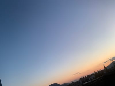 This media file is a photograph of a sky over a town in Khust, Ukraine. The subjects are the sky, clouds, a few buildings with chimneys, and rolling hills. The scene depicts a town in the distance as the sun sets or rises.
The time of day is evident from the gradient of colors in the sky, transitioning from a deep blue at the top to lighter shades of blue, orange, and yellow near the horizon. This indicates either sunrise or sunset. There are a few scattered clouds in the sky, some appearing to catch the light of the sun.
The setting appears to be a residential area with buildings that have traditional-looking chimneys, suggesting a somewhat older or European architectural style. The rolling hills in the background frame the town, providing a natural element to the scene.
There are no people or specific activities or events clearly depicted. The overall emotion evoked is one of tranquility and the beauty of a natural transition of light.
There is no visible text in the image.