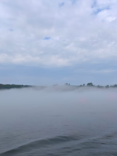 The image depicts a serene natural landscape, likely a body of water, shrouded in a thick layer of fog. The foreground features the textured surface of the water, with gentle ripples suggesting calm conditions. Beyond the water, a dense mist obscures the middle ground, creating a hazy, ethereal effect. In the distance, the faint outline of a tree line and what appears to be a structure, possibly a building with a white roof, can be discerned through the fog. The sky above is overcast, with soft, diffused light filtering through the clouds. The scene is tranquil and evokes a sense of quietude.