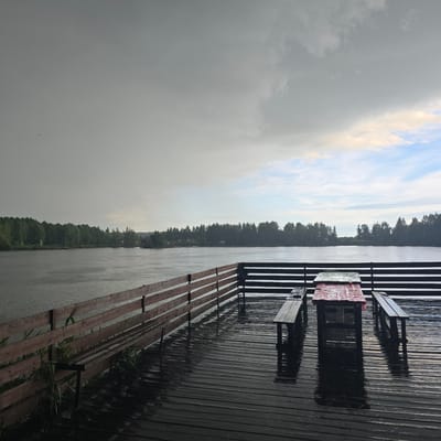 This image depicts a wet, wooden deck overlooking a body of water, likely a lake or river, with a dense forest lining the opposite bank. The sky is overcast with some lighter patches indicating the possibility of clearing weather. Rain is visible in the foreground on the deck and the water's surface. On the deck, there is a picnic table covered with a red and white checkered plastic cloth, and two benches. A wooden railing runs along the edge of the deck, separating it from the water. The overall atmosphere is somber and wet, suggesting recent or ongoing rain. The scene is set in Дубровка, Russia, as indicated by the provided location context.
