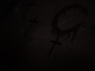 The image depicts a dark, unlit environment with faint markings on a textured surface. Three distinct crosses are visible, two of which appear to be drawn or painted. One cross is located in the upper left quadrant, and another is positioned in the lower center. A larger, more abstract symbol resembling a stylized 'C' or a broken circle with drips is present in the upper right quadrant. The texture of the surface suggests it could be concrete, stone, or a similar rough material. The lighting is extremely dim, with significant areas of shadow obscuring detail. No people or specific objects other than the markings are discernible. The context of Gukovo, Russia, is not visually evident from the image itself.