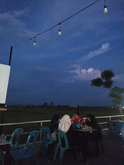 In Cotcut, Indonesia, at dusk, several people are seated at a table in an outdoor dining area. The sky is a deep blue, with a few wispy clouds and a string of bare light bulbs strung across it. A white projection screen is visible on the left. The ground is covered in dark grass, and a fence runs along the edge of the dining area. In the background, distant lights suggest a populated area. The overall atmosphere is relaxed, with people enjoying a meal or drinks as the day transitions into night.