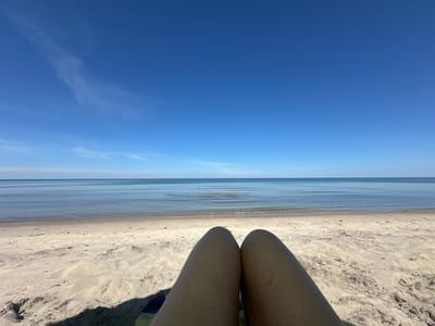The image depicts a serene beach scene in Lesnoy, Russia, under a clear blue sky. The foreground is dominated by a person's legs, tanned and resting on what appears to be a beach towel or mat, casting a shadow on the sand. The sand is light-colored and textured, with scattered debris like small stones and twigs. Beyond the sand, a calm, expansive body of water stretches to the horizon, its surface rippled and reflecting the sky. A thin line of surf breaks gently along the shoreline, separating the sand from the water. The overall atmosphere is tranquil, suggesting a leisurely day at the beach.