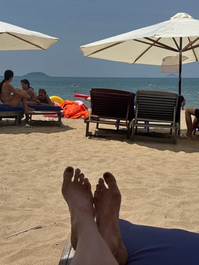 The image captures a beach scene in Nha Trang, Vietnam. In the foreground, a pair of feet rests on a blue beach lounger, with sand covering the skin and toenails. The sand stretches out to a line of beach chairs and umbrellas. To the left, two women in bikinis recline on loungers. Further back, a group of people are seen by the water, some with inflatable rings. The ocean is calm, with a distant island visible on the horizon under a clear blue sky. The scene appears to be a relaxed day at the beach.