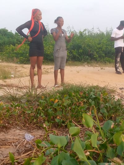 Two women and a man stand on a sandy beach in front of thick vegetation under an overcast sky. The woman on the left wears a short black dress with orange braids and is posed with her hands on her hips. The woman to her right wears a grey dress and gestures with her hands. A man in a white shirt and black pants stands to the right of the second woman. Close to the foreground, lush green vegetation with broad leaves stretches across the bottom of the frame. A piece of litter sits in the sand amongst the leaves.