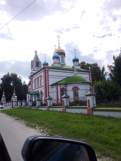 The image depicts the Church of the Intercession of the Most Holy Theotokos in Staryi Pokrov, Russia. The church, a prominent religious structure, features white walls with red trim, several blue and golden domes topped with crosses, and a green-tiled roof. It is surrounded by a white and red brick fence with an ornate black metal railing. Lush green grass and trees border the church and the adjacent road. The scene is captured from a low angle, possibly from a moving vehicle, as indicated by the presence of a car's rearview mirror in the foreground. The sky is partly cloudy, suggesting overcast weather conditions. No people are visible in the frame.