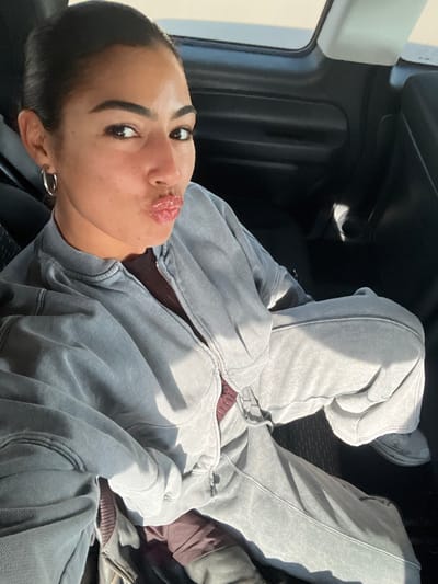 The scene is located within the interior of a vehicle, in Deerlijk, Belgium. A female subject, appearing to be an adult with dark hair pulled back, brown eyes, and a light to medium skin tone, is seated in what appears to be a rear passenger seat. She is looking directly at the camera, posing with her lips pursed in a "kiss" gesture. She wears a light gray, faded-wash, full-zip hooded sweatshirt over a dark maroon ribbed top, and matching light gray, faded-wash sweatpants. A silver-toned hoop earring is visible on her left ear. The vehicle's interior features dark upholstery and dark plastic door panels. A visible sunroof or window opening at the top of the frame indicates daylight, with direct sunlight illuminating the subject and portions of the vehicle interior, creating distinct highlights.