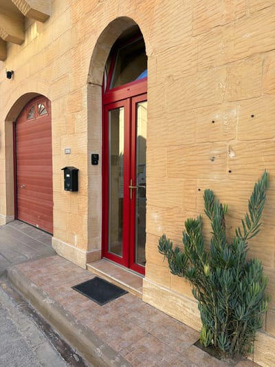 This is a medium shot, eye-level exterior photograph of a building's entrance in Xagħra, Malta. The building facade is constructed of large, tan-colored stone blocks with a rough, horizontal texture. To the left, a red garage door is visible, set within an arched opening. To the right of the garage door, on the stone wall, is a small black mailbox and a small black doorbell or intercom device. A silver house number '41' is also affixed to the wall above the mailbox. Adjacent to the garage door is a main entrance with a double red door and a glass transom. The door is framed by a large stone arch. A small black doormat sits on the tiled walkway in front of the double doors. To the right of the entrance, a green succulent plant grows from a small planter box in the tiled walkway. The overall lighting suggests it is daytime, possibly late afternoon, with shadows indicating the sun's position. The ground in front of the walkway is a grey, paved surface.