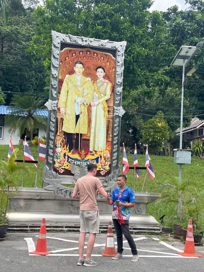 In a scene set in Tambon Wang Prachan, Thailand, a large framed portrait of a royal couple is prominently displayed. The portrait is ornate, with a decorative silver frame featuring intricate scrollwork. The couple is depicted in traditional, formal attire, with the male figure wearing a golden robe and the female figure in a matching golden outfit.
Two men are visible near the portrait. One man, wearing a blue shirt with a colorful abstract design and black pants, stands to the left, looking towards the portrait. He is holding some red and white streamers. The other man, wearing a light brown t-shirt and grey shorts, is on the right, facing away from the camera and towards the portrait. He appears to be adjusting something on the portrait or its frame.
The setting appears to be outdoors, possibly along a street or pathway, indicated by the asphalt ground and white lines. Several orange traffic cones are placed along the edge of the road. In the background, there is lush green foliage from trees and palm fronds. A light pole with a solar panel is also visible. Several small Thai flags are affixed to a pole to the left of the portrait. The overall lighting suggests it is daytime, with overcast conditions or the shade of trees obscuring direct sunlight. The atmosphere seems casual, with the men engaged in activity related to the portrait. No specific event is clearly indicated, but the display of the royal portrait suggests a public or ceremonial space.