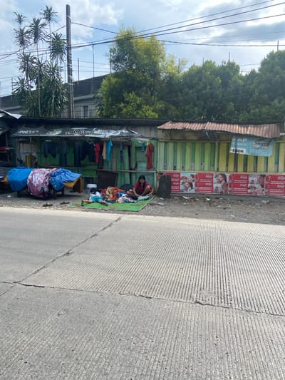 The scene is located in Kecamatan Dayeuhkolot, Indonesia, at the coordinates -6.965 latitude and 107.615 longitude. There are two women sitting on a mat in front of a concrete building with a green corrugated metal wall. One woman is wearing a red shirt and the other is wearing a blue shirt. A young child is lying on their stomach in front of the women on the mat. The women and the child are facing the camera, and the child is looking toward the left of the frame. The building is made of concrete and wood and has a roof of corrugated metal. There is a sign on the wall, and it appears to have a logo for a company named "Siera". The building is surrounded by vegetation, including trees and shrubs. The concrete road in the foreground is cracked and has grooves on the surface. There is a pile of clothes, a plastic container, and a small amount of debris scattered on the ground. The sky is partly cloudy. The scene appears to be a residential area.