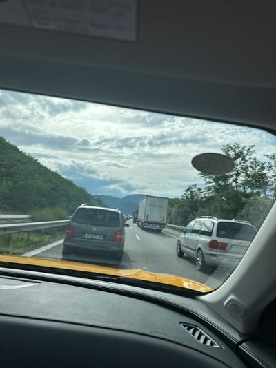 The image shows a view from inside a vehicle, looking out through the windshield at a traffic jam on a highway. The road is lined with guardrails and slopes upward to a forested hillside on the left. Several vehicles are visible in traffic, including a gray Volkswagen Touran, a white panel van, and a silver station wagon with a Bulgarian license plate. The sky is cloudy, with patches of blue peeking through. The scene is consistent with a highway in or near Gorno Varshilo, Bulgaria, given the presence of Bulgarian license plates and the mountainous, forested landscape typical of the region.