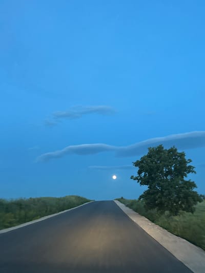 The image depicts a road scene at twilight in Smilets, Bulgaria. The sky is a light blue with wispy clouds. A full moon is visible near a tree with green foliage. Adjacent to the tree is a paved road with grassy verges on both sides. The road surface is dark and reflects light. The image is rotated.