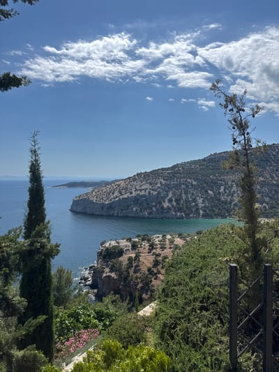 The image displays a scenic coastal view from a vantage point overlooking the sea and a rugged, tree-covered hillside. In the foreground, lush green foliage and a tall, slender evergreen tree frame the left side of the composition. A dark metal fence is visible on the right.

The middle ground features a dramatic cliff face descending to the water. The sea is a deep blue, transitioning to a turquoise hue in a sheltered cove. The hillside is densely populated with scrubby trees and shrubs, with a winding path visible in the distance.

In the background, a broad expanse of the sea stretches to the horizon, where a faint outline of distant islands can be seen. The sky is a bright blue, adorned with scattered white, wispy clouds.

The scene is consistent with a location such as Moni Archaggelou, Greece, known for its natural beauty and coastal landscapes. There are no people or active events depicted in the image. The overall impression is one of tranquility and natural grandeur.