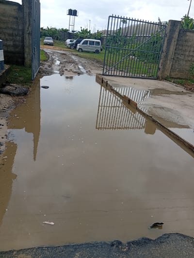 The image depicts a waterlogged entrance area in Igrita, Nigeria. A large puddle of muddy water dominates the foreground, reflecting the gate, walls, and sky above. The gate itself is partially open, with metal bars and concrete pillars on either side. Beyond the gate, a dirt road shows signs of disrepair with potholes and puddles. Several vehicles, including a white van and a silver car, are parked further down the road. In the background, a water tower is visible. The sky is overcast, suggesting recent rain. A discarded tire and various pieces of debris are visible in the muddy area.