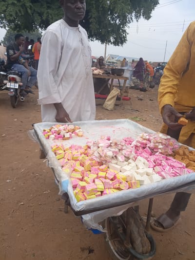 In Tilde, Nigeria, a vendor stands behind a cart filled with colorful confectionery. The cart is laden with small, cube-shaped candies, predominantly pink, yellow, and white, some individually wrapped in plastic. The vendor, wearing a white robe, has his right hand near the candies, suggesting he may be arranging or serving them. To the right, another person in a yellow garment holds a piece of candy. In the background, a bustling marketplace is visible with people, motorcycles, and stalls. The ground is dirt, and trees with sparse foliage are present, indicating an outdoor setting. The overall scene depicts a vibrant market environment where local goods are being sold.