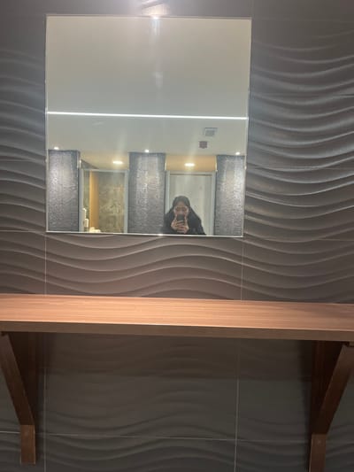 The image captures a contemporary interior space, most likely a bathroom or dressing area, with a strong focus on reflective surfaces and textured walls. A large, rectangular mirror dominates the left side of the frame. The wall surrounding the mirror is adorned with dark gray or black tiles that feature a prominent, undulating wave-like texture, arranged vertically. To the right, a light-colored wooden structure, potentially part of a vanity or shelving unit, runs vertically against the textured wall.
Reflected prominently in the mirror is a person, presumably the photographer, taking a mirror selfie with a smartphone held up to their face. The person has dark hair and is seen wearing a dark garment, with a discernible slight smile. The reflection also reveals the interior of the room opposite the mirror: a brightly lit space with white walls and horizontal strip lighting. Within this reflected area, two dark, possibly sparkling or textured, decorative panels frame another section that appears to be an opening or a recessed area. Further back in the reflection, details of a light-tiled space, possibly a shower or bath area, with what seem to be decorative candles, can be glimpsed. Small, indistinct colored squares are also visible on the reflected white wall.
The activity taking place is the act of capturing a mirror selfie. The overall mood conveyed by the person's slight smile is positive or neutral. The lighting is artificial, suggesting an indoor setting where time of day is not explicitly conveyed. No discernible text is visible in the image. The scene suggests a modern, well-maintained, and stylish environment.