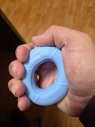 A person's hand is gripping a light blue, circular hand grip strengthener. The grip is textured on the inner and outer edges and has white Cyrillic lettering on the upper outer surface that reads "ЖМИ PRO". The background is a blurred wooden surface. The image was taken in Anadyr, Russia.