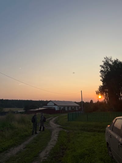 The image depicts a rural scene at sunset in the village of Perebor, Russia. Two individuals in camouflage uniforms are standing on a dirt path in a grassy area. Behind them, a white house with a red roof is visible, bordered by a green wooden fence. To the right of the scene, the sun is setting behind trees, casting a warm glow. A portion of a dark-colored car is parked on the right side of the frame, with its rear end facing the viewer. The sky exhibits a gradient of colors, transitioning from a pale blue at the top to orange and pink hues near the horizon. There are utility poles and wires present in the middle ground. The overall environment suggests a peaceful, rural setting during dusk.