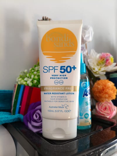 The image displays a collection of cosmetic and personal care products arranged on a black tray, likely in a bathroom or dressing area. The primary subject is a large white tube of "Bondi Sands SPF 50+ Very High Protection Fragrance Free Water Resistant Lotion." The tube is prominently featured, with text indicating its sun protection factor, benefits like "72hr hydration," and suitability for "sensitive skin." Other visible items include a blue bottle with the brand name "Ambre Solaire" and a "Ready to Go" indicator suggesting it's a travel-sized product. Several decorative items, such as artificial flowers in shades of pink, orange, and white, and a purple fabric rose, add a touch of color and texture to the scene. The background is blurred, with hints of white and soft lighting, suggesting an indoor setting. The overall impression is that of a personal care product display, possibly showcasing items for sun protection and skincare. There are no people visible in the image. The scene appears to be indoors, with artificial lighting. There are no specific cues to suggest the time of day, weather, or precise location within Sandwell, United Kingdom.