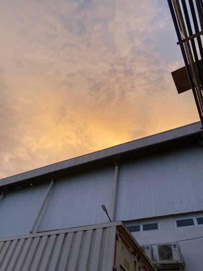 The image is an upward-angle shot of a building's corrugated metal roof and a cloudy sky, taken in Gambut Barat, Indonesia. The sky is filled with soft, diffused clouds, illuminated with a warm, golden-orange hue, suggesting either sunrise or sunset. The building's roof is a prominent feature, its corrugated texture creating a repetitive pattern of light and shadow.
On the right side of the image, the side of another building is visible, featuring several rectangular windows with white frames and dark panes. Below these windows, an outdoor air conditioning unit is mounted on the building's exterior. Further to the right, a section of a white container or truck is visible, with some red and yellow markings on its surface. A light fixture with a rectangular head extends from the roofline of the main building.
The scene appears to be outdoors, likely in an industrial or commercial area, given the building materials and the presence of a container. The time of day is indicated by the dramatic lighting of the sky, pointing towards twilight. The overall mood is calm and contemplative, with the warm sky contrasting with the industrial structures. No people are visible in the image. There is no discernible text in the image.