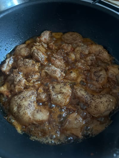 The image shows pieces of chicken being cooked in a dark non-stick frying pan. The chicken appears to be seasoned with black pepper and possibly paprika, and is simmering in a rich, golden-brown sauce. The pan is partially filled with the sauce, and small bubbles are visible around the chicken, indicating it is actively cooking. The background is out of focus and shows the edge of what appears to be a stovetop or kitchen counter. There is no indication of a specific location in the city of Peremyshliany, Ukraine.