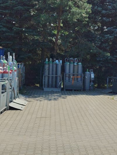 The image shows an outdoor storage area for industrial gas cylinders. In the foreground, a paved area with interlocking concrete blocks extends towards the background. To the left, a silver metal rack holds several gas cylinders, some with red and white labels, and others with green caps. Two metal ramps are visible on the ground in front of this rack.
In the middle ground, two more racks of gas cylinders are present. The left rack contains several grey cylinders with green caps, held in place by a dark crate-like structure with an orange strap. To the right of this rack, a single grey cylinder with a green cap stands next to a smaller grey cylinder with a white label, which is itself stacked on another green-capped cylinder. In the background, a dark metal fence runs along the edge of the property, behind which tall evergreen trees create a dense backdrop. The lighting suggests it is daytime with bright sunlight, casting shadows on the paved area. The scene appears to be a functional storage area, likely at an industrial facility or a gas supplier's yard in Palmiry, Poland. No people are visible in the image.