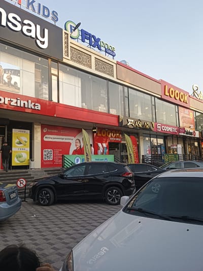 The image shows a street scene in front of a shopping complex in Yangiiul, Uzbekistan. The complex features prominent signage for various businesses, including "KIDS," "nsay," "FIX price GROUP," "Korzinka," "LOOOK," "ASLAR," and "COSMETIC." Several cars are parked on the patterned pavement in front of the complex. A person in a black t-shirt and grey pants is walking away from the Korzinka entrance, which has a display of corn and cabbage for sale. A red sign with a white "R" inside a red circle indicates a no-parking zone. The sky is clear and blue, suggesting it is daytime. The architecture of the shopping complex includes large glass windows and red and beige facade elements.