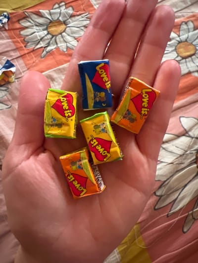 A hand is holding five individually wrapped candies, all with the "Love is..." branding and featuring a red heart. The candies are a mix of yellow and orange wrappers, with some green trim. The background is a pink and orange fabric with a daisy pattern. The image is a close-up shot, with the hand and candies occupying the majority of the frame. The lighting is natural and appears to be from a window. The location of Новокатаево, Russia is not discernible from the image.