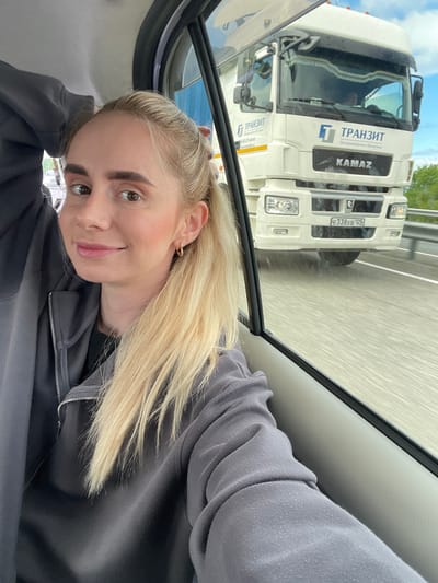 A young blonde Caucasian woman, positioned in the lower-left to mid-frame, is shown from the chest up, taking a selfie inside a vehicle. She has fair skin, dark eyebrows, and brown eyes looking directly at the camera with a subtle smile. Her long blonde hair is gathered in a ponytail, flowing over her right shoulder. She wears a dark gray hooded zip-up sweatshirt over a black garment, and a small golden hoop earring is visible in her right ear. Her left hand is partially visible near the back of her head.
Through the vehicle's window on the right, a white KAMAZ brand semi-trailer truck is visible, driving on a highway. The truck features the "ТРАНЗИТ" (TRANSIT) logo and text in Cyrillic on its side and grille. Its front displays a Russian license plate, "E338XB 125," indicating the Primorsky Krai region. The road surface below the truck is blurred, suggesting motion. A metal guardrail parallels the highway, and green trees are visible in the background under a partly cloudy sky. The scene depicts travel on a highway in the region of De-Friz, Primorsky Krai, Russia.