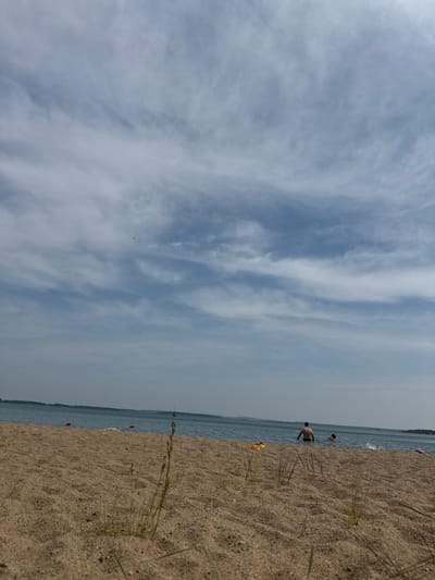 The image depicts a serene beach scene with sandy shores meeting a calm body of water. The sky is a pale blue, filled with wispy, elongated clouds. In the distance, a faint tree line is visible along the horizon.

In the water, two individuals are present. One person, a male with tanned skin, stands partially submerged, facing away from the camera. Another individual, possibly a child, is further out in the water, with only their head and shoulders visible. The water shows some ripples and splash activity around the second individual.

On the sandy beach, there are a few scattered objects: a pair of yellow flip-flops are positioned on the sand, and a pink object, possibly a float or towel, is visible to the right. Several red buoys are dotted in the water, suggesting a designated swimming area or marker.

The foreground is dominated by coarse sand, with sparse blades of dry grass growing from it. The perspective is low, making the sand texture prominent. The overall impression is that of a quiet, recreational moment on a warm day. This scene is located on the shore of Lake Aiyrtau, a significant natural landmark within the city of Aiyrtau, Kazakhstan, known for its recreational beaches and waters.