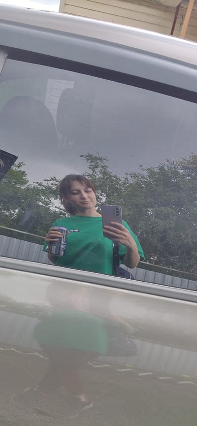 A woman is reflected in a vehicle window. She is wearing a green t-shirt and holding a can of drink and a smartphone. The reflection shows trees and a fence in the background. The woman appears to be taking a selfie. The environment seems to be outdoors, possibly in a parking lot or on a street in Tashkazgan, Russia.