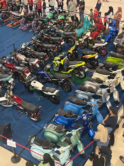 The image depicts an indoor exhibition or event, likely a motorcycle show, set up on a blue carpeted floor. The exhibition features a diverse collection of motorbikes and scooters, predominantly in vibrant colors such as red, blue, green, yellow, and white. The vehicles are arranged in rows, with some elevated on stands. Several people are visible in the background and foreground, observing the display. Some are standing and looking at the vehicles, while others appear to be in conversation. The environment suggests a public space, possibly a mall or convention center, with what looks like temporary barriers and signage. Based on the provided context of Dumai City, Indonesia, this scene is likely taking place in a public venue within the city.