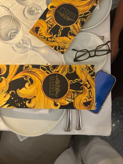 The image captures a dining table scene at Gianni's at The Former Versace Mansion, located in Miami Beach, United States. The table is draped with a white tablecloth. Two menus for "GIANNI'S AT THE FORMER VERSACE MANSION" are visible, featuring a black background with elaborate gold baroque patterns and chains. White ceramic plates are positioned on the tablecloth, accompanied by clear glassware, including a water glass and partially visible wine glasses. A pair of dark-framed reading glasses rests on one of the white plates. A smartphone with a blue screen/case is partially visible on the table. Silverware is arranged alongside the plates. A person's leg, clad in white trousers, is partially visible at the bottom of the frame, indicating an occupant at the table. A partial arm is also visible on the right side of the frame.