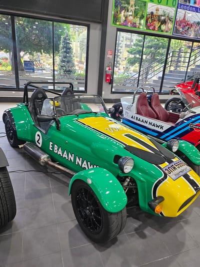 The image shows a bright green, open-top sports car with a yellow stripe, parked indoors on a gray tiled floor. The car has a roll cage, two seats, and the number "2" on its side. Text on the car reads "E BAAN NAWK" and "NONG NOOCH PATTAYA". A license plate on the front reads "3กพ 5555". Behind the green car, another sports car with red seats and "AI BAAN NAWK" text is visible, followed by a red motorcycle. Large windows in the background reveal a garden setting with many cacti and lush greenery, and a sign pointing to "Cafe Amazon". The overall environment appears to be a showroom or a visitor center.