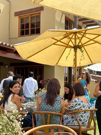 The image shows an outdoor gathering of people at a table. The location appears to be La Molina, Peru, based on the architectural style. Several individuals are seated at the table, engaged in conversation or looking down at the table. The table is decorated with flowers and glassware. The scene is partly shaded by large yellow patio umbrellas, which give off a bright glow around the edges. Buildings are visible in the background, and the image is taken from an elevated perspective, looking down on the scene.