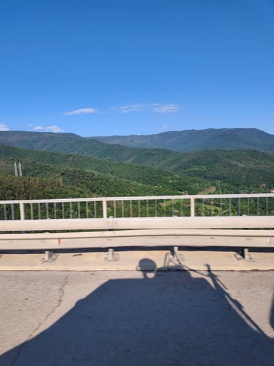 A sunny day over a lush, green mountain range in Vrachesh, Bulgaria is depicted from a bridge. The foreground features a concrete road surface with distinct shadows of people cast on it, suggesting movement. A white metal guardrail runs horizontally across the mid-ground, separating the road from the expansive vista. Beyond the guardrail, rolling hills covered in dense green forest stretch into the distance, with a clear blue sky dotted with a few wispy clouds overhead. A single, tall electricity pylon is visible on one of the forested slopes. The overall impression is one of natural beauty and a bright, clear day in a mountainous region.