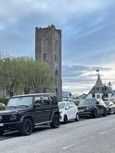 The image depicts a street scene in Reykjavik, Iceland, with Hallgrímskirkja church prominently visible in the background. A row of parked cars lines the right side of the street. A black Mercedes-Benz G-Class SUV is parked closest to the viewer. A tree is in front of the church. The sky is overcast with light grey and white clouds. Other buildings are seen in the background, adding to the urban landscape of the city.