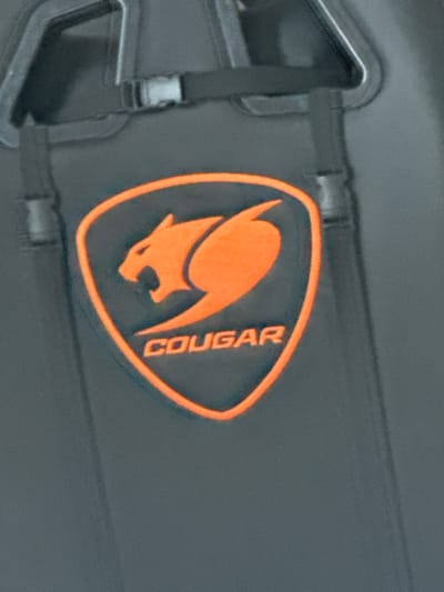 The image presents a close-up, static view of a dark grey, slightly textured surface, likely fabric or molded plastic. Centrally positioned is an embossed or embroidered orange logo for the brand "COUGAR." The logo features a stylized cougar or mountain lion in an aggressive, pouncing stance with its mouth open, enclosed within an orange shield-shaped outline. Below the animal graphic, the brand name "COUGAR" is rendered in orange, capitalized block letters. Above the logo, dark grey webbing straps are visible. One strap runs horizontally across the top, featuring a small, light grey plastic side-release buckle. Another dark grey strap appears vertically, connecting to a larger, dark grey, curved or U-shaped structural element at the top of the frame. No individuals, specific actions, or environmental cues are visible to ascertain the precise nature of the object or its surrounding location. The image does not contain any information to indicate a location in Saraia, Bulgaria, or any other specific geographical area.