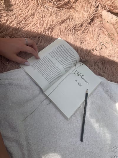 An overhead shot shows an open book lying on a white textured towel, with a pink fuzzy blanket in the background. A person's hand, with long fingernails painted light pink, rests on the left page of the book. A silver ring is visible on their index finger. A dark gray pencil with a pattern of small stars lies on the right page of the book, which has a chapter heading that reads "Часть вторая" in Russian. A few blades of dry grass are scattered on the blanket and towel. The scene appears to be set outdoors, possibly in a park or backyard. The lighting suggests it is daytime, with sunlight casting shadows. The image does not contain any specific elements that would indicate its location within the city of Пліскі сельскі Савет, Belarus.
