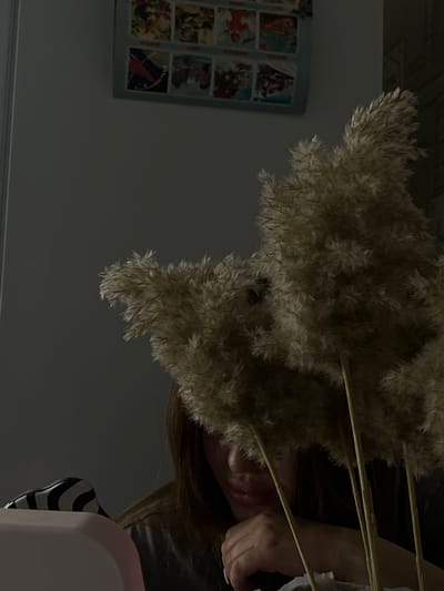 The image shows a close-up view of a person partially obscured by a bunch of dried pampas grass. The person appears to be holding the bunch in front of their face. Only a portion of their face, including the nose and lips, is visible. Their hair is long and brown. Behind the person and the grass is a wall with a framed poster or picture of multiple small images. To the lower right, part of a pink object with a black and white striped element is visible. The setting appears to be indoors, likely a home. There is no information to locate the scene in the city of Port-Katon, Russia.